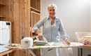 Eine freundliche Frau bügelt in einem hellen Raum. Im Hintergrund sind ein Wäschekorb und eine Waschmaschine zu sehen.