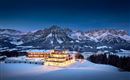 Ein modernes Hotel in einer schneebedeckten Landschaft mit beeindruckenden Bergen im Hintergrund. Die Fassade des Hotels leuchtet warm im Dämmerlicht.