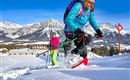 Zwei Frauen wandern im Schnee mit Schneeschuhen in einer malerischen Berglandschaft. Die Sonne scheint und die Berge sind schneebedeckt.