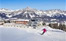 Eine verschneite Berglandschaft mit einem modernen Hotel und majestätischen Bergen im Hintergrund. Im Vordergrund fährt ein Skifahrer die Piste hinunter.