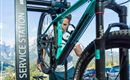 A person is working on a bicycle at a service station. In the background, mountains and cars can be seen.