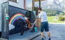 A man is washing a bicycle at a wash station with a rainbow design. In the background, mountains and a green landscape can be seen.