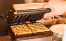 Een wafelijzer met verse, goudbruine wafels wordt geopend. Op de achtergrond zijn servies en ingrediënten zichtbaar.