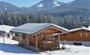 A cozy wooden cabin in the snow, surrounded by snow-covered mountains and forests. The clear sky and winter landscape create a peaceful atmosphere.