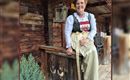 A smiling woman is sitting on a wooden rock in front of a rustic cabin. She is wearing a traditional outfit and has short, reddish-brown hair.