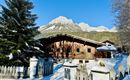A cozy wooden house in the snow with snow-covered mountains in the background. The surroundings are characterized by fir trees and a white fence.