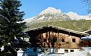 A cozy wooden house in the snow with a wooden protective wall. In the background, majestic mountains rise up under a clear blue sky.