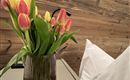 Váza s barevnými tulipány stojí na stole. V pozadí je vidět dřevěná zeď a polštář.