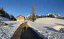 A snowy landscape with a large wooden house and snow-covered mountains in the background. The path leads through the idyllic winter scenery.