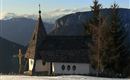A small church in a snowy landscape with mountains in the background. The sky is clear and blue.