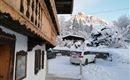 A winter landscape with snow-covered trees and mountains in the background. A traditional wooden house is visible in the foreground, next to a car parked in the snow.