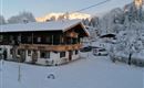 A charming, wintry farmhouse surrounded by snow. The mountains in the background are coated with snow, and the atmosphere is calm and picturesque.