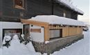 A cozy wooden house with a snow-covered roof in a wintry landscape. The entrance area is simply and invitingly designed.