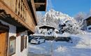 A traditional wooden house in the snow, surrounded by a wintry landscape. In the background, snow-covered mountains and a clear blue sky can be seen.
