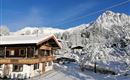 A cozy wooden house in a snowy landscape. In the background, a majestic mountain range rises under a clear blue sky.