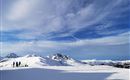 A winter landscape with snow-covered mountains and a clear sky. In the foreground, some people can be seen skiing or hiking.