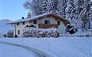 A cozy house in the snow, surrounded by tall, snow-covered fir trees. The path gently leads to the front door.