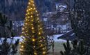 Ein schöner, beleuchteter Weihnachtsbaum steht in einer winterlichen Landschaft. Im Hintergrund sind schneebedeckte Berge und Tannen zu sehen.