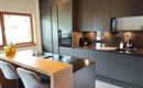 A modern kitchen with dark cabinets and an elegant countertop. At the table, there are chairs, and daylight is coming in through the window.