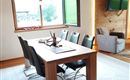 A modern dining room with a wooden table and black chairs. Large windows offer a view of the landscape.