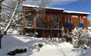 Ein modernes Holzhaus im Schnee, umgeben von winterlicher Landschaft. Der Himmel ist klar und blau, und die Bäume sind mit Schnee bedeckt.