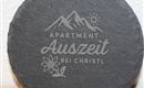 Ein runder, grauer Stein mit der Aufschrift "Apartment Auszeit bei Christl". Darüber sind stilisierte Berge und eine Sonne sowie eine Blume abgebildet.