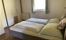 Ein helles Schlafzimmer mit einem Doppelbett und einem großen Fenster. Der Raum hat einen schlichten Stil mit Holzfußboden und einem Kleiderschrank.