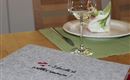 Ein schöner Tisch gedeckt mit einem Glas Wein und einem dekorierten Teller. Auf dem Tisch liegt eine graue Stoffmappe mit dem Schriftzug „Willkommen!“.
