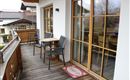 Ein gemütlicher Balkon mit Holzdielen, zwei Stühlen und einem Tisch. Durch die großen Fenster ist ein schöner Blick nach draußen möglich.
