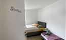 Een lichte kamer met twee bedden en eenvoudige inrichting. Er is een smalle ruimte met een houten vloer en eenvoudige decoratie.
