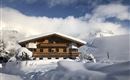 A cozy wooden house in the snow, surrounded by a wintry landscape. The mountains are visible in the background, and the sky is partly cloudy.