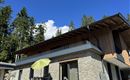 Een modern huis met een steen- en houten gevel, omringd door bomen. Op het terras staan tafels en stoelen, evenals een gele parasol.