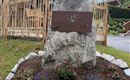 A large rock with a metal plaque displaying the name "Auen." Colorful flowers and stones decorate the area around the rock.