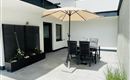 A modern terrace with a large sun umbrella and a dining area. In the background, plants and a simple wall can be seen.