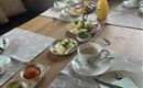 Ein schön gedeckter Frühstückstisch mit Tassen, Geschirr, Marmelade, Käse und frischem Obst. Im Hintergrund sind Brötchen und ein Glas Saft zu sehen.