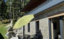Een terras met een tafel en stoelen, overdekt door een groene parasol. Op de achtergrond zijn bomen en een gezellig huis te zien.