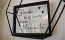 Ein gerahmtes Glasbild mit der Aufschrift: „Glück ist die Summe schöner Momente.“. Der Rahmen ist schwarz und hat eine moderne Form.