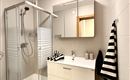 A modern bathroom with a shower and a sink. The decor is bright and minimalist, featuring a large mirror and striped towels.