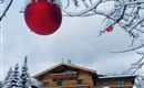 Ein schneebedecktes Landschaftsbild mit einem hübschen Holzhaus. Im Vordergrund hängt eine rote Weihnachtskugel an einem Baum.