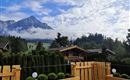 Eine malerische Berglandschaft mit hohen Gipfeln und bewölktem Himmel. Im Vordergrund eine Holzterrasse mit grünen Pflanzen und Blick auf angrenzende Häuser.