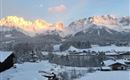 A wintry landscape with snow-covered mountains in the background. Small houses are visible nearby, while the sun illuminates the mountain peaks.