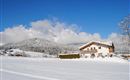 A snowy landscape with a charming house and majestic mountains in the background. The sky is clear and blue, creating a serene winter atmosphere.