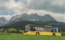 A yellow bus drives through a green meadow with majestic mountains in the background. The sky is cloudy, which gives the scene a peaceful atmosphere.