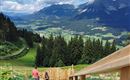 Eine malerische Berglandschaft mit grünen Wiesen und dichten Wäldern. Im Vordergrund sind Wanderer auf einem Holzweg zu sehen.