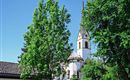 A church with a tall tower, surrounded by green trees. The sky is clear and blue.
