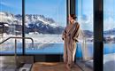 Eine Frau in einem Bademantel steht am Fenster und blickt auf die verschneiten Berge. Im Vordergrund ist ein Schwimmbad sichtbar, das die wunderschöne Aussicht zeigt.