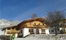 A typical alpine house with wooden cladding in a snowy environment. In the background, the mountains and a clear sky can be seen.