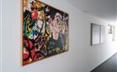 Ein farbenfrohes Kunstwerk hängt an einer weißen Wand im Flur. Im Hintergrund sind weitere Bilder und ein Fenster sichtbar.