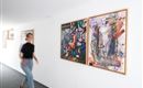 Eine Person geht an bunten Gemälden entlang, die an einer weißen Wand hängen. Der Raum hat ein modernes und helles Ambiente.