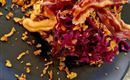 Een bord met paarse coleslaw, knapperige spekjes en gebakken uien. Het gerecht ligt op een zwart bord en is decoratief opgediend.
